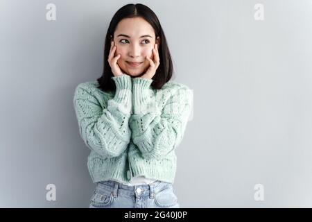 La ragazza pensiva tiene il viso con le mani. Bruna carina in un maglione lavorato a maglia pensando a qualcosa su uno sfondo di una parete grigia. Foto Stock