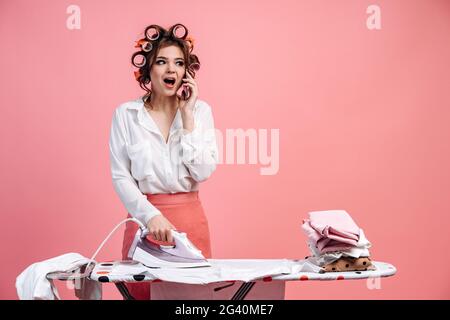 Sorpresa casalinga ragazza con arricciatori sulla sua testa ferri una montagna di vestiti, parla al telefono ed è molto sorpreso per qualche ragione. Bella casa Foto Stock