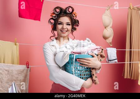 Giovane casalinga con i arricciacapelli sulla testa che tiene un cesto con i vestiti. Sullo sfondo di lacci appesi vestiti su una corda su una parete rosa. Foto Stock