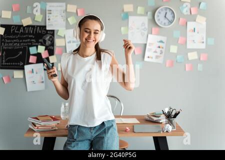 Una ragazza positiva studentessa in cuffie si trova nella sua stanza vicino al desktop, su uno sfondo grigio con adesivi colorati. La ragazza ha divertimento, ascolta musi Foto Stock