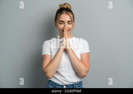 Una giovane donna in una T-shirt bianca casual prega, aggrappando le mani, chiudendo gli occhi. Giovane ragazza isolata su sfondo grigio. Foto Stock