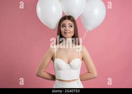 Eccitata, gioiosa ragazza fa una sorpresa, nasconde palloncini dietro la schiena. Carino signora e palloncini su uno sfondo rosa, eccitato, gioiosa ragazza fa un s. Foto Stock