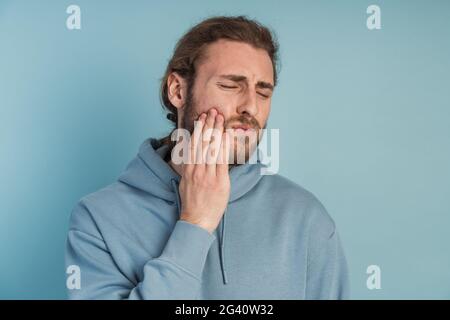 Dolore ai denti. Giovane uomo che sente dolore, che tiene la guancia con entrambe le mani, che soffre di mal di denti, che ha un'espressione dolorosa Foto Stock