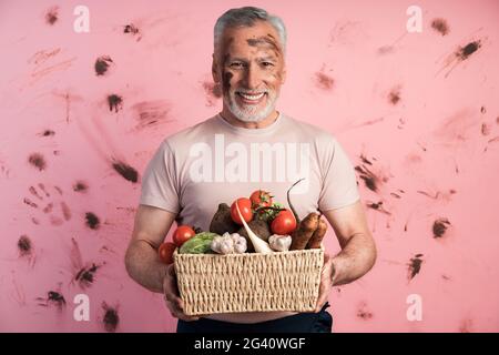 Uomo anziano sorridente e positivo che tiene un cesto di verdure fresche contro una parete rosa sporca. Il concetto di agricoltura, casa., sorridente, positivo sen Foto Stock