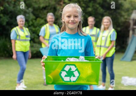 Caucasica ragazza che tiene scatola di riciclaggio con gruppo di uomini e. donne che raccolgono rifiuti nel campo dietro Foto Stock
