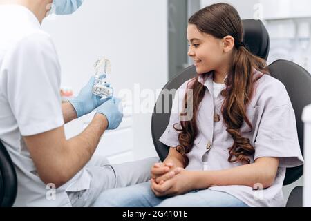 Il dentista comunica con il paziente, mostra sulla mascella come spazzolare i denti correttamente. La ragazza è un adolescente in una sedia dentale. Foto Stock