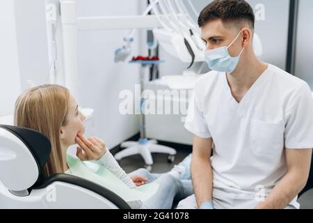 Il dentista in una maschera protettiva comunica con un paziente attraente. Il dentista lavora nell'ufficio odontoiatrico Foto Stock