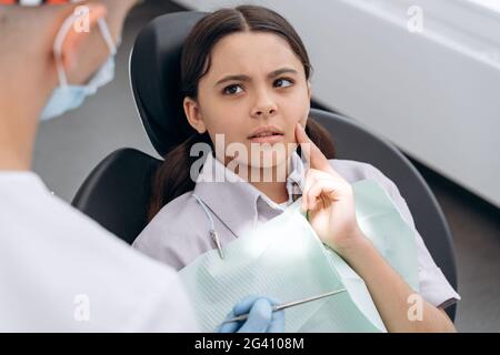 La ragazza teen triste tocca le guance con le dita, si lamenta di mal di denti. Il dentista comunica con un paziente giovane Foto Stock