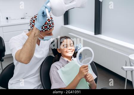 Il dentista comunica con un paziente giovane. La ragazza in una sedia dentale tiene uno specchio, insieme con il medico esamina i denti Foto Stock