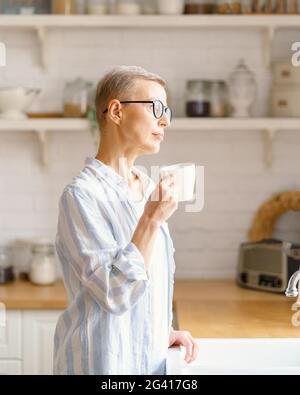 Bella donna anziana con tazza di caffè o acqua rure guardando in finestra mentre godendo di nuovo giorno a casa, verticale Foto Stock