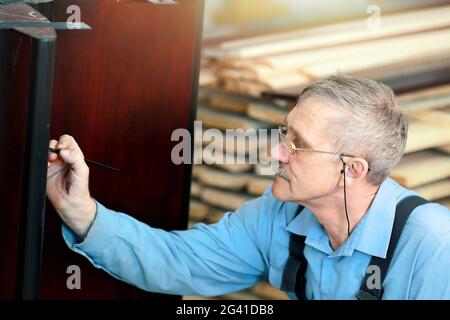 Un uomo anziano con occhiali e capelli grigi lavora con legno in un negozio di carpenteria. Foto Stock