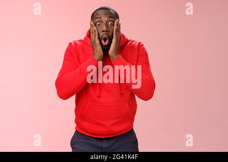 WOW incredibile. Ritratto impressionato sorpreso afroamericano bearded ragazzo in rosso hoodie pieghevole labbra piegando occhi allargati scioccato Foto Stock