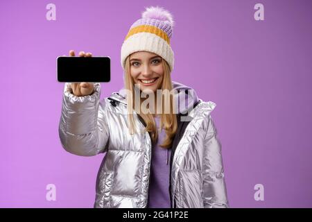 Ragazza bionda sicura e amichevole all'aperto con giacca glittering argento cappello tenere smartphone orizzontale mostrando pho mobile Foto Stock
