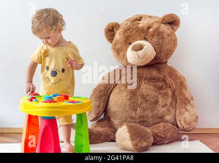 Bambino dolce felice che si diverte a giocare in un tavolo colorato di attività con il suo enorme orsacchiotto, al coperto a casa Foto Stock