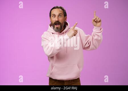Impressionato scioccato scosso fresco hipster maturo 50s uomo bearded capelli grigi in rosa felpa con cappuccio retelling incredibile storia che punta in su Foto Stock