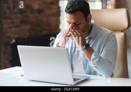 L'uomo indiano stanco che usa il computer portatile per lavorare, un ragazzo in abbigliamento casual tocca e massaggiare il ponte del naso, si sente stanco e scoppiato, seduto alla scrivania in un ufficio moderno con gli occhi chiusi Foto Stock