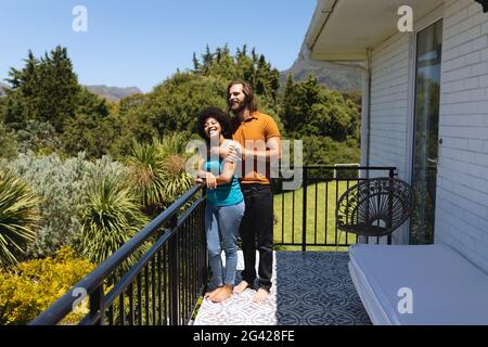 Felice coppia diversa in piedi sul balcone abbracciando e sorridendo Foto Stock