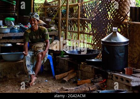 Un cuoco balinese, seduto in una cucina rampicante in una capanna, fumando e ridendo. Bali, Indonesia. Foto Stock