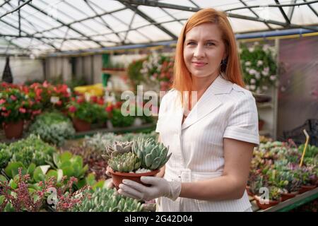 Ritratto di donna rossastra indossando guanti di gomma e vestiti bianchi che tengono succulenti o cactus in pentole con altre piante verdi sullo sfondo Foto Stock
