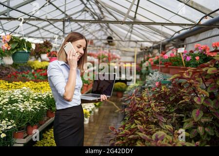 Sorridente proprietario della serra che posa con un laptop nelle sue mani che parla sul telefono che ha molti fiori in background e tetto di vetro. Spazio per il testo sullo schermo. Foto Stock