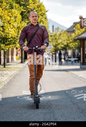 Giovane uomo in pantaloni e camicia, borsa sopra la spalla, guidando scooter elettrico verso la fotocamera Foto Stock