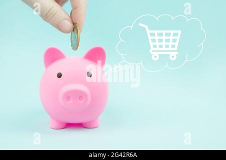 Mano che mette una moneta in una banca piggy con carrello di shopping ologramma digitale in nube pensato sopra la sua testa su sfondo blu. Risparmio di denaro, finanziario p Foto Stock