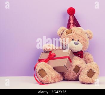 Carino orsacchiotto marrone in un cappuccio rosso si siede e tiene una scatola marrone con un regalo, sfondo festoso Foto Stock
