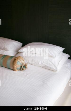 Un letto con una coperta bianca e cuscini di diverse dimensioni e un cuscino allungato in una camera con pareti scure. Foto Stock