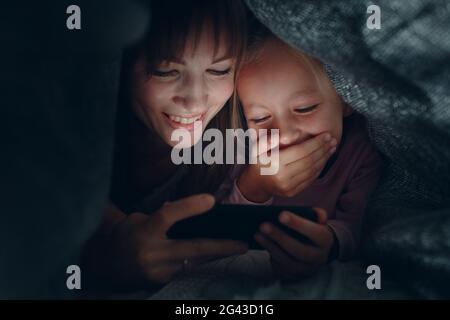 Madre con una piccola figlia che guarda i contenuti sullo smartphone al buio sotto le coperte Foto Stock