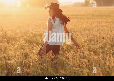 Donna contadina in cappello cowboy che cammina con le mani sulle orecchie al campo agricolo al tramonto Foto Stock