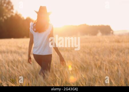 Donna contadina in cappello cowboy che cammina con le mani sulle orecchie a campo di grano agricolo al tramonto Foto Stock