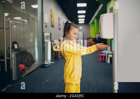 Bambina che usa l'erogatore automatico di gel di alcool spruzzando sulle mani disinfettante antisettico macchina igienizzante, nuova vita normale af Foto Stock