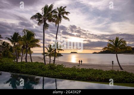 Piscina, alberi di cocco e una coppia a piedi sulla spiaggia al Six Senses Fiji Resort al tramonto, Malolo Island, Mamanuca Group, Isole Fiji, Sud Foto Stock