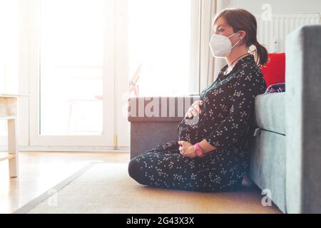 Quarantena a casa. Donna incinta con maschera ffp2 è seduta sul pavimento nel soggiorno Foto Stock