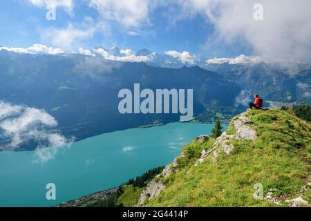 Donna mentre camminando siede sulla sporgenza dei prati e si affaccia sul Lago di Brienz e sulle Alpi Bernesi sullo sfondo, da Augstmatthorn, Alpi Emmentali, Berna Foto Stock