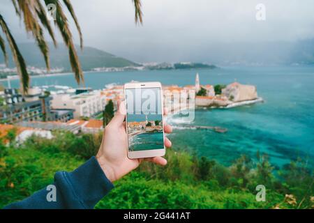 La mano dell'uomo tiene uno smartphone con una foto di architettura sullo sfondo della città vecchia di Budva, Montenegro Foto Stock