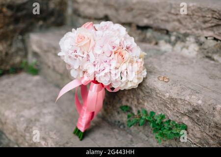 Bouquet nuziale di rose rosa e idrangea, con nastri rosa giù sui gradini di pietra e anelli di nozze vicino ad esso Foto Stock