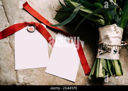 Gli anelli di nozze legati con i nastri rossi giacciono sulle schede vuote con un ornamento accanto ad un mazzo di fiori su uno sfondo di to Foto Stock