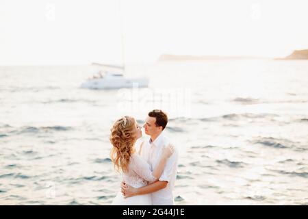 La sposa e lo sposo stanno abbracciando sulla riva e stanno per baciare, uno yacht bianco veleggia dietro di loro Foto Stock