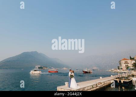 La sposa e lo sposo si abbracciano sul molo vicino alla città vecchia di Perast, le barche galleggiano nelle vicinanze Foto Stock