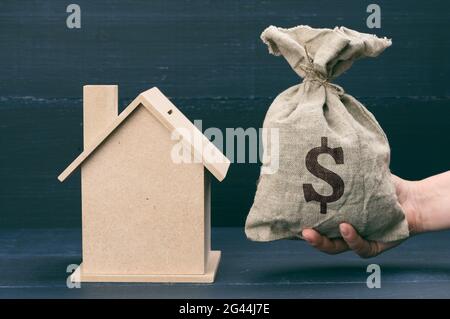 La mano tiene una borsa di tela piena di soldi e una casa di legno su uno sfondo blu. Il concetto di acquisto del bene immobile, locazione fuori Foto Stock