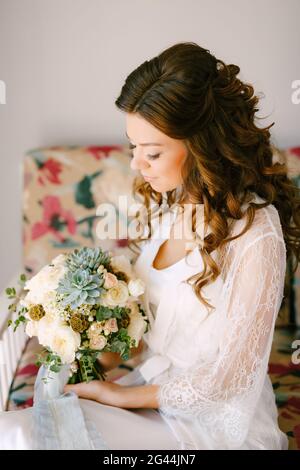 Una sposa in un abito di pizzo si siede su un divano in una camera d'albergo e tiene un bouquet di nozze nelle sue mani, da vicino Foto Stock