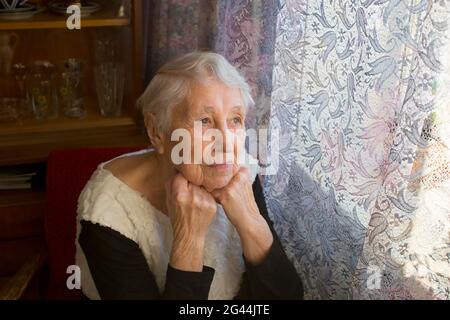 Vecchia donna sola seduta vicino alla finestra di casa sua e guardando e sognando Foto Stock