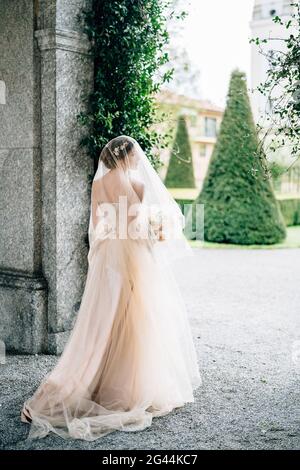 Sposa in un abito da sposa con un velo e un bouquet di fiori si trova vicino all'arco di un vecchio edificio di fronte a un giardino verde. L Foto Stock