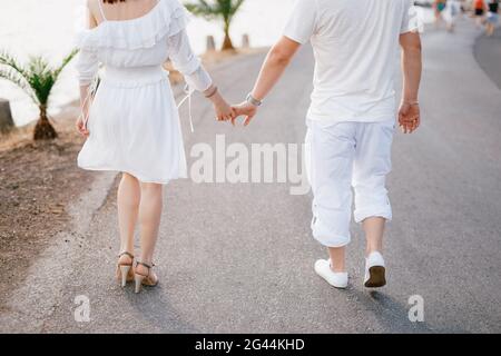 Un paio di amanti in abiti bianchi camminano lungo la strada vicino al mare, una donna tiene delicatamente la mano di un uomo Foto Stock