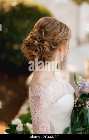 Una sposa con una bella acconciatura in un abito in pizzo si erge con un bouquet e guarda a lato, primo piano Foto Stock