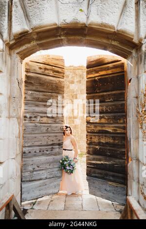 Bella sposa in un bel vestito con un bouquet di i fiori si appoggiano contro la porta di legno aperta del castello Foto Stock