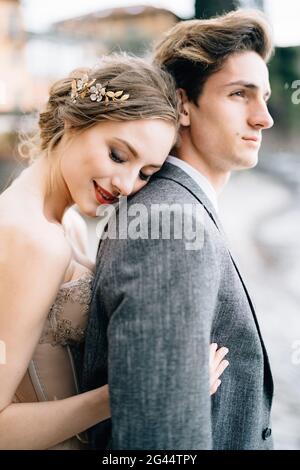 Sorridente sposa abbracca lo sposo da dietro mentre si trova sulla passeggiata del Lago di Como. Mezzo ritratto Foto Stock