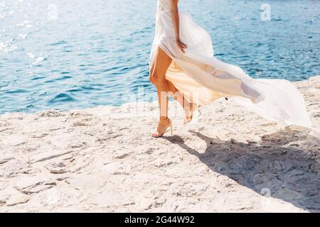 Le gambe della sposa in sandali dai tacchi alti e con una gonna che si gonna in fluttering, la sposa cammina lungo il molo, da vicino Foto Stock