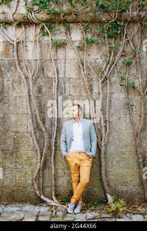 Lo sposo pensivo in un vestito di colore luminoso si leva contro un muro di pietra intrecciato con edera verde Foto Stock